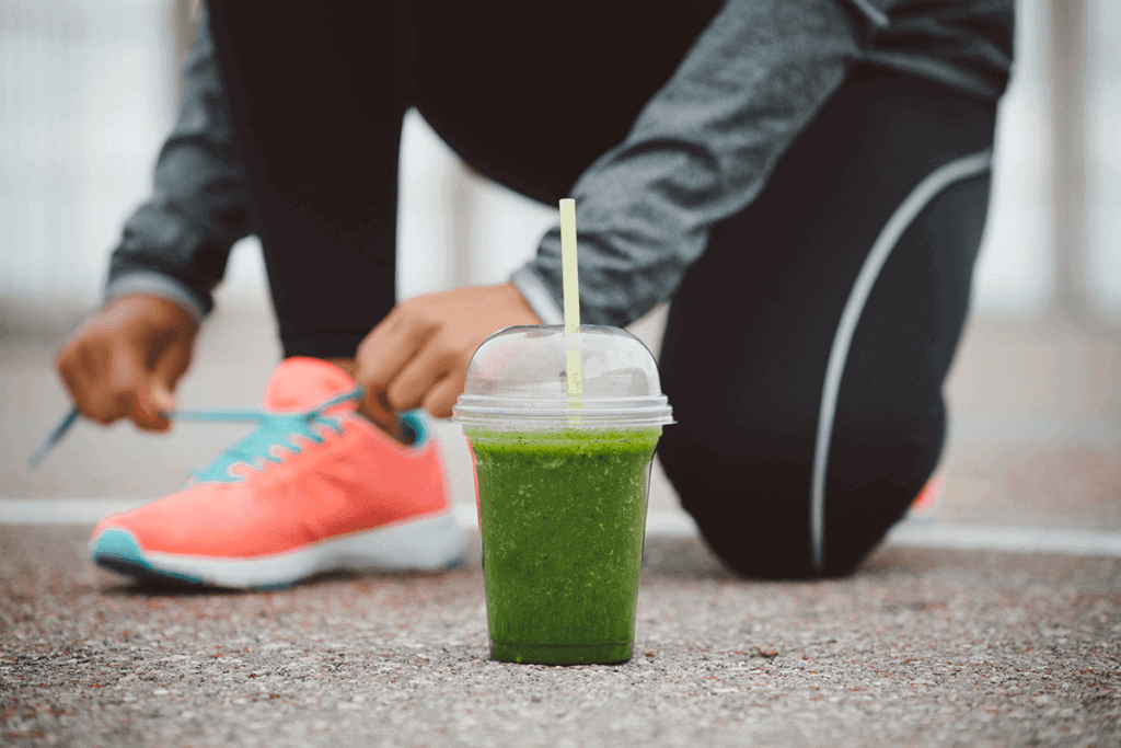 close up of nutritious green drink on the ground with woman tying her running shoe in the background