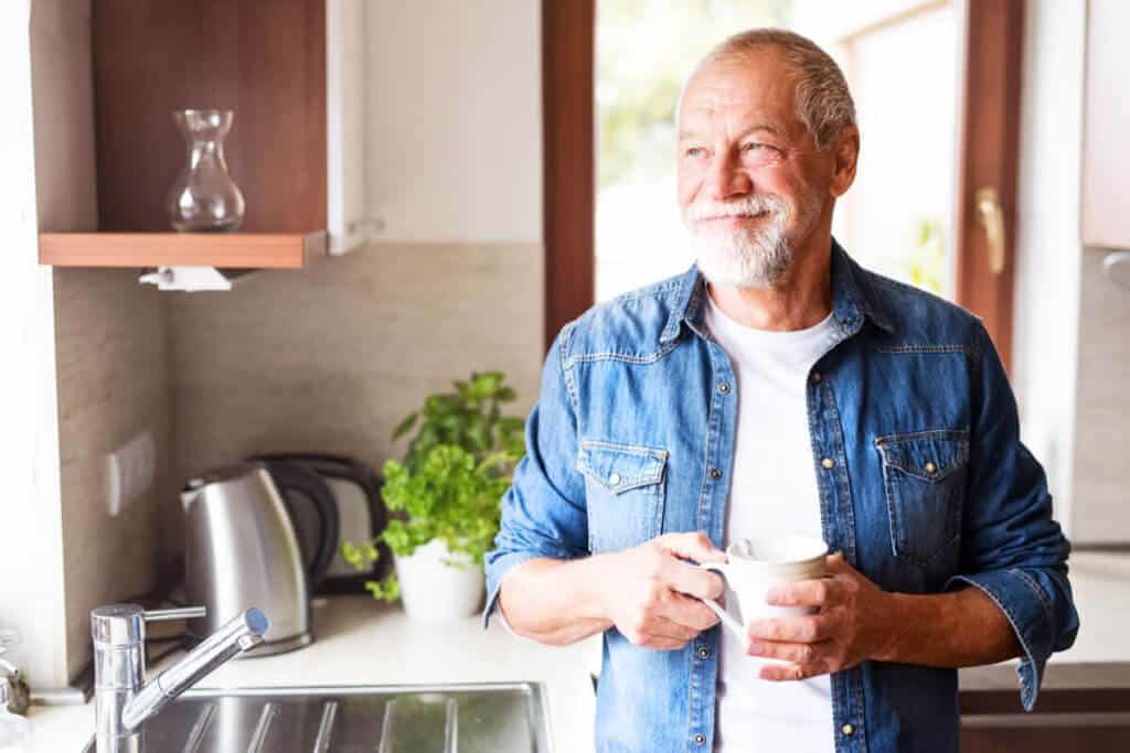 older man standing in kitchen looking out the window and smiling holding a mug