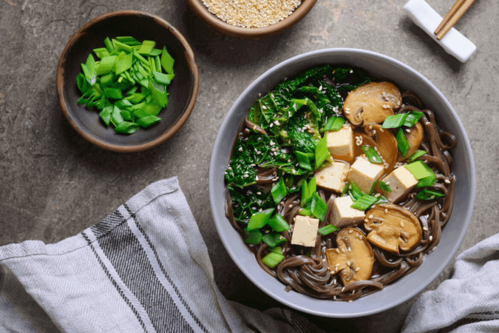 A bowl of Asian noodles with mushrooms, tofu, and greens on a grey backdrop.