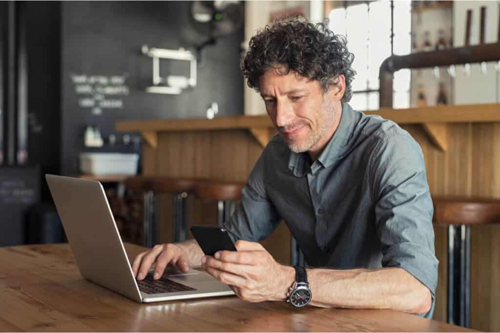 man sitting in coffee shop looking at phone and working on laptop