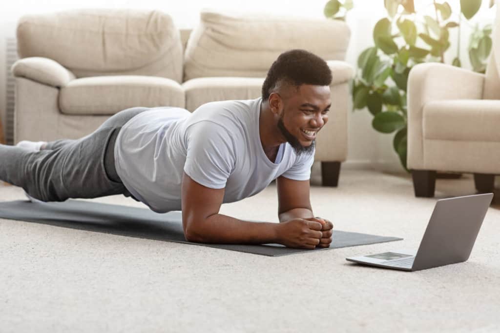 person planking in front of a laptop on a mat in their living room