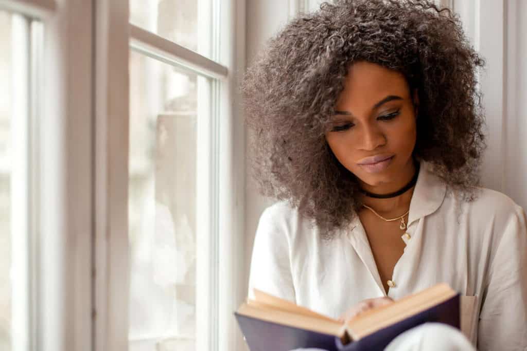 African American woman sitting on windowsill reading a book.