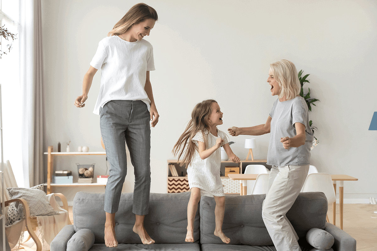 grandmother, daughter, and granddaughter jump on a grey couch together