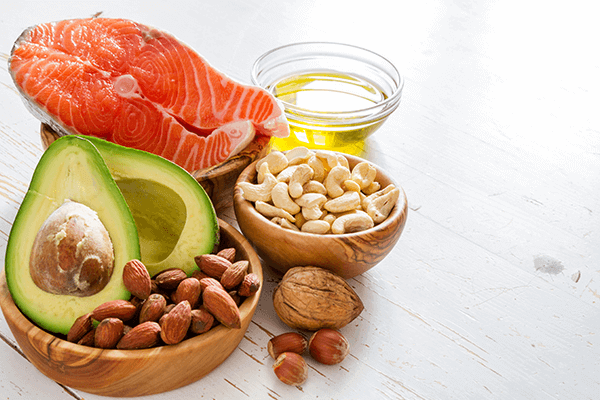 clear glass filled with oil, wooden bowl filled with cashews, wooden bowl filled with almonds and a cut up avocado, and wooden bowl filled with a piece of meat